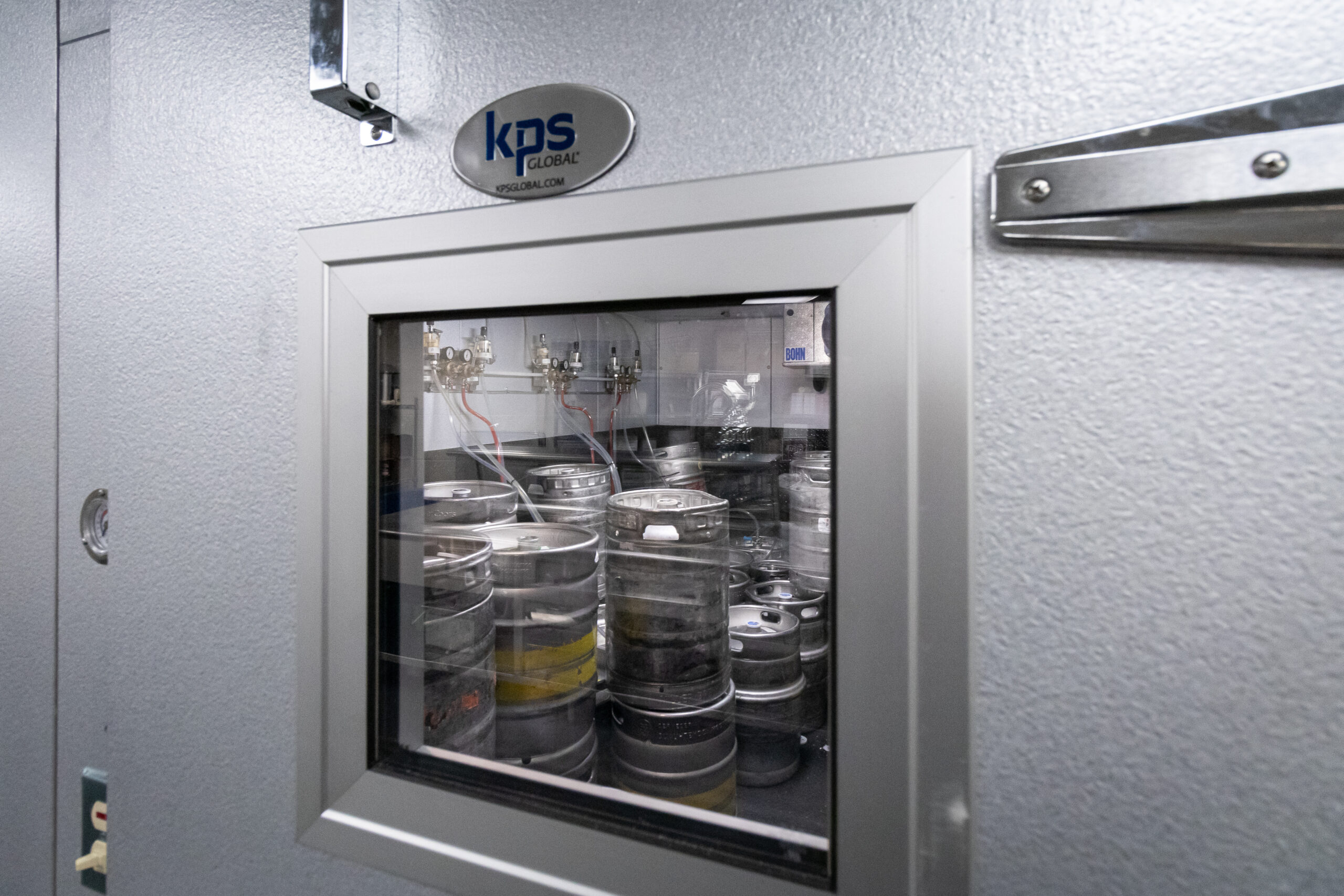 View through the glass door of a walk-in beer cooler, revealing stacked beer kegs